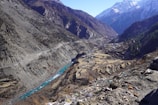 A mountainous landscape featuring steep, rocky slopes and a narrow valley with a meandering turquoise river running through it. On one side of the valley, terraced fields and a small settlement with a few structures are visible. Snow-capped peaks tower in the background, providing a dramatic backdrop. The foreground includes scattered debris and litter.