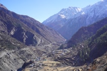 A scenic valley surrounded by towering mountains with a mix of rocky and forested slopes. Snow-capped peaks are visible in the distance under a clear blue sky. A small village with scattered houses and terraced fields is situated along the valley floor, connected by winding roads.