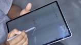 A person holds a digital pen and writes 'close' on a tablet screen. The hand is gripping the stylus, while the tablet displays a drawing or note-taking app. The background is blurred, indicating focus on the device.