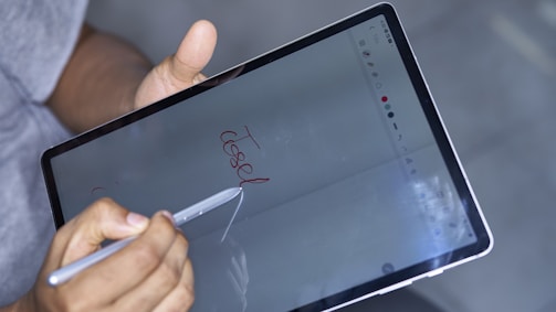 A person holds a digital pen and writes 'close' on a tablet screen. The hand is gripping the stylus, while the tablet displays a drawing or note-taking app. The background is blurred, indicating focus on the device.