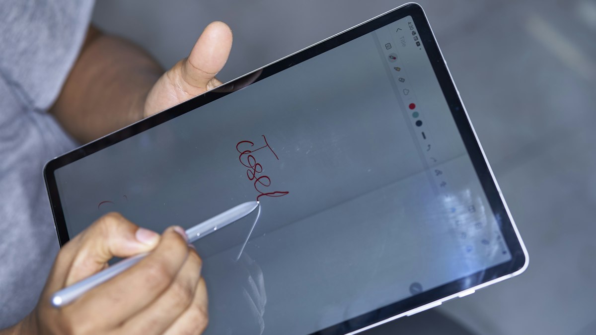 Hand signing a name in red ink on a tablet screen with a stylus