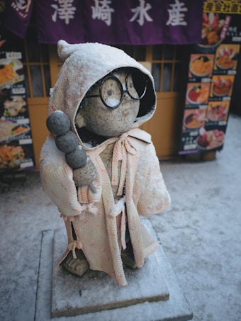 A stone statue dressed in a textured robe and hood, adorned with round glasses, stands in a snowy environment. The statue holds a skewer with round objects and is situated in front of a building with purple curtains featuring white characters. Menus with food images are displayed beside the statue.