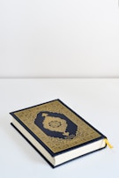 A book with an ornate gold and dark blue cover featuring intricate patterns and Arabic calligraphy lies on a flat white surface. A yellow ribbon bookmark peeks out from the pages.