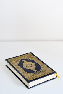 A book with an ornate gold and dark blue cover featuring intricate patterns and Arabic calligraphy lies on a flat white surface. A yellow ribbon bookmark peeks out from the pages.
