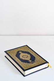 A book with an ornate gold and dark blue cover featuring intricate patterns and Arabic calligraphy lies on a flat white surface. A yellow ribbon bookmark peeks out from the pages.