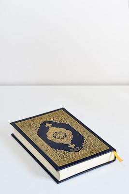 A book with an ornate gold and dark blue cover featuring intricate patterns and Arabic calligraphy lies on a flat white surface. A yellow ribbon bookmark peeks out from the pages.