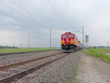 A bright red and yellow locomotive travels on a set of parallel railway tracks, set amidst a rural landscape. The overcast sky forms a drab background, with power lines and pylons visible in the distance. The railway is bordered by gravel and stretches into the horizon, surrounded by green fields.