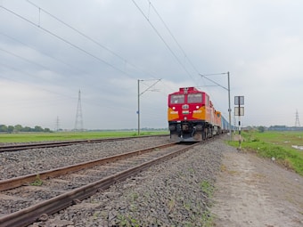 A bright red and yellow locomotive travels on a set of parallel railway tracks, set amidst a rural landscape. The overcast sky forms a drab background, with power lines and pylons visible in the distance. The railway is bordered by gravel and stretches into the horizon, surrounded by green fields.