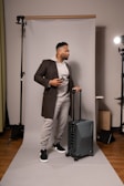 A man stands on a photography backdrop holding a smartphone and a suitcase. He is wearing a brown coat over a gray hoodie and sweatpants, with black and white sneakers. The setting includes professional lighting equipment.
