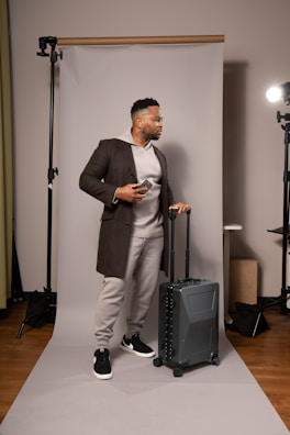A man stands on a photography backdrop holding a smartphone and a suitcase. He is wearing a brown coat over a gray hoodie and sweatpants, with black and white sneakers. The setting includes professional lighting equipment.