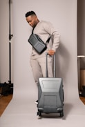 A person in a neutral-toned outfit is standing on a plain backdrop, holding a suitcase with a sleek design. They have a crossbody bag and are wearing matching sweatpants and a sweatshirt. The environment appears to be a studio setup.
