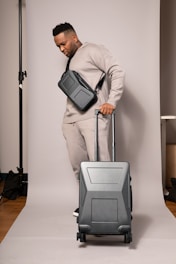 A person in a neutral-toned outfit is standing on a plain backdrop, holding a suitcase with a sleek design. They have a crossbody bag and are wearing matching sweatpants and a sweatshirt. The environment appears to be a studio setup.
