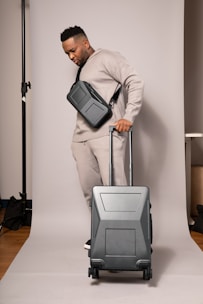 A person in a neutral-toned outfit is standing on a plain backdrop, holding a suitcase with a sleek design. They have a crossbody bag and are wearing matching sweatpants and a sweatshirt. The environment appears to be a studio setup.