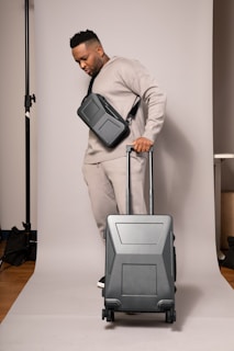 A person in a neutral-toned outfit is standing on a plain backdrop, holding a suitcase with a sleek design. They have a crossbody bag and are wearing matching sweatpants and a sweatshirt. The environment appears to be a studio setup.