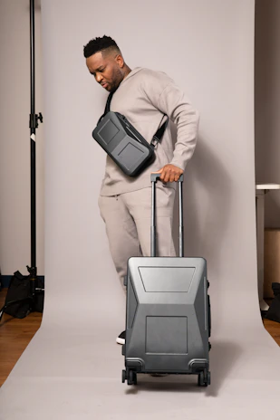 A person in a neutral-toned outfit is standing on a plain backdrop, holding a suitcase with a sleek design. They have a crossbody bag and are wearing matching sweatpants and a sweatshirt. The environment appears to be a studio setup.