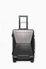 A sleek, modern hardshell suitcase with a dark matte finish, featuring a telescopic handle and four wheels. The suitcase has a minimalistic design with geometric patterns and appears sturdy and durable.
