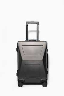 A sleek, modern hardshell suitcase with a dark matte finish, featuring a telescopic handle and four wheels. The suitcase has a minimalistic design with geometric patterns and appears sturdy and durable.