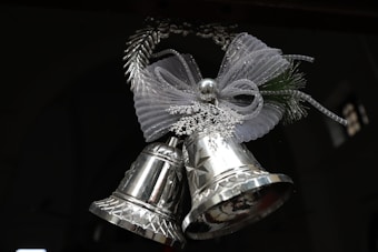 A pair of shiny silver bells adorned with an intricate ribbon bow made of mesh and decorative snowflakes. The bells hang in a festive arrangement suggesting a holiday theme.