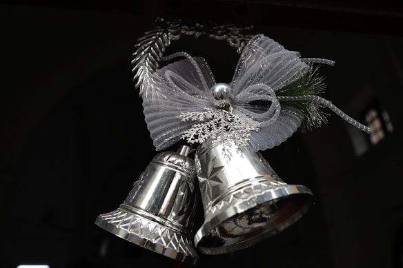 A pair of shiny silver bells adorned with an intricate ribbon bow made of mesh and decorative snowflakes. The bells hang in a festive arrangement suggesting a holiday theme.