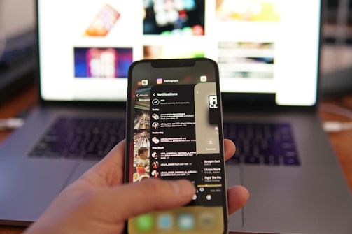 A person holds a smartphone displaying Instagram notifications, with a focus on the screen. A blurred laptop is visible in the background, suggesting multitasking or technology use.