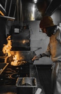 A chef in a beanie and glasses is actively cooking in a professional kitchen, holding a pan with flames rising high. The kitchen appears industrial, with stainless steel equipment and dim lighting.