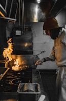 A chef in a beanie and glasses is actively cooking in a professional kitchen, holding a pan with flames rising high. The kitchen appears industrial, with stainless steel equipment and dim lighting.