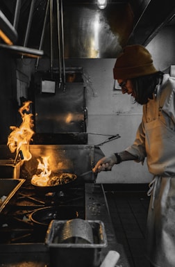 A chef in a beanie and glasses is actively cooking in a professional kitchen, holding a pan with flames rising high. The kitchen appears industrial, with stainless steel equipment and dim lighting.