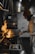 A chef in a beanie and glasses is actively cooking in a professional kitchen, holding a pan with flames rising high. The kitchen appears industrial, with stainless steel equipment and dim lighting.