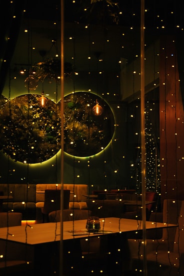 A dimly lit interior setting with warm, ambient lighting. Small, glowing fairy lights are strung across the view, casting a cozy and intimate atmosphere. Circular wall decorations with greenery are visible, alongside a hanging lamp. Plush seating and a simple wooden table complete the inviting arrangement.