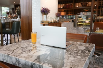 A modern cafe setting with a marble table featuring a white laptop and a glass of orange juice. The background includes warm lighting, brown wooden accents, shelves with various items, and a bouquet of dried flowers.