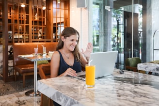 A woman sitting at a marble table in a cafe, engaging in a video call on a laptop. She is smiling and waving, with a glass of orange juice placed on the table. The cafe interior includes a mix of wooden and modern decor, with a few tables and chairs visible in the background.