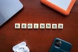 Scrabble tiles spelling the word 'BEGINNER' are arranged on a wooden surface surrounded by technological items, including a smartphone, a laptop, an external hard drive, and wireless earphones.