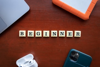 Scrabble tiles spelling the word 'BEGINNER' are arranged on a wooden surface surrounded by technological items, including a smartphone, a laptop, an external hard drive, and wireless earphones.