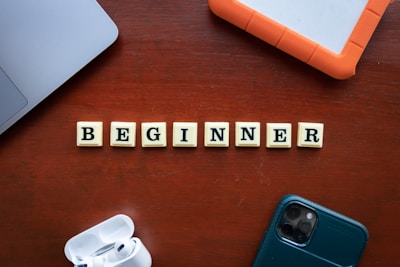Scrabble tiles spelling the word 'BEGINNER' are arranged on a wooden surface surrounded by technological items, including a smartphone, a laptop, an external hard drive, and wireless earphones.