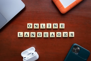 Wooden tiles with letters spelling out 'ONLINE LANGUAGES' are arranged neatly on a wooden surface. Surrounding the tiles, there are a laptop, a hard drive, a smartphone, and wireless earphones.