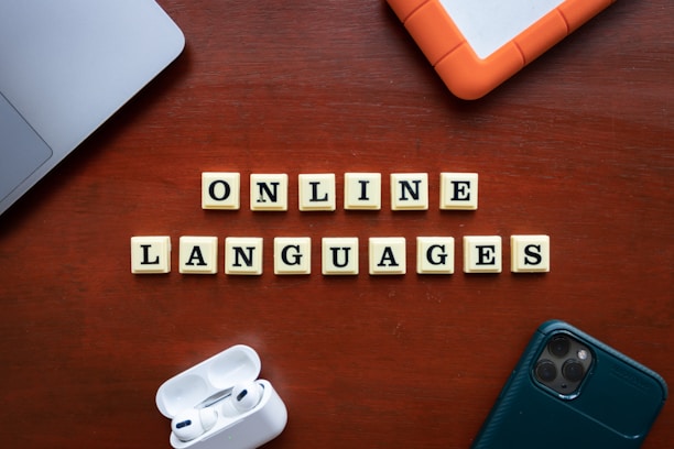 Wooden tiles with letters spelling out 'ONLINE LANGUAGES' are arranged neatly on a wooden surface. Surrounding the tiles, there are a laptop, a hard drive, a smartphone, and wireless earphones.