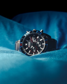 A sleek and modern wristwatch with a black dial and metallic strap is prominently placed against a backdrop of luminous blue fabric. The watch face features multiple sub-dials and red accents, giving it a sophisticated and sporty appearance.