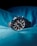 A sleek and modern wristwatch with a black dial and metallic strap is prominently placed against a backdrop of luminous blue fabric. The watch face features multiple sub-dials and red accents, giving it a sophisticated and sporty appearance.