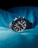A sleek and modern wristwatch with a black dial and metallic strap is prominently placed against a backdrop of luminous blue fabric. The watch face features multiple sub-dials and red accents, giving it a sophisticated and sporty appearance.