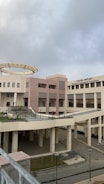 A modern architectural building with a circular structure on top and Arabic and English text on its facade, indicating it is the College of Languages & Translation. The building has a light beige and pink color scheme with large windows and external corridors. The sky appears overcast, casting a muted light over the scene.