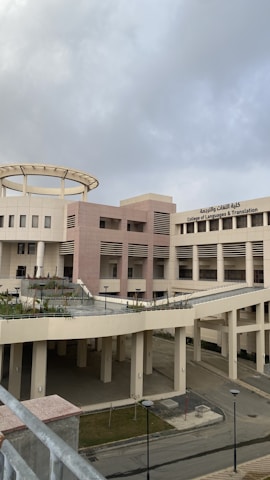 A modern architectural building with a circular structure on top and Arabic and English text on its facade, indicating it is the College of Languages & Translation. The building has a light beige and pink color scheme with large windows and external corridors. The sky appears overcast, casting a muted light over the scene.