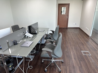 A modern office workspace with a row of desks equipped with dividers and ergonomic chairs. Items such as laptops, a phone, and stationery are neatly placed on the glass-topped desks. The office is minimally decorated, with wooden flooring and neutral-colored walls. A closed wooden door is focused in the background, and a floor-to-ceiling glass panel is seen on the right side.