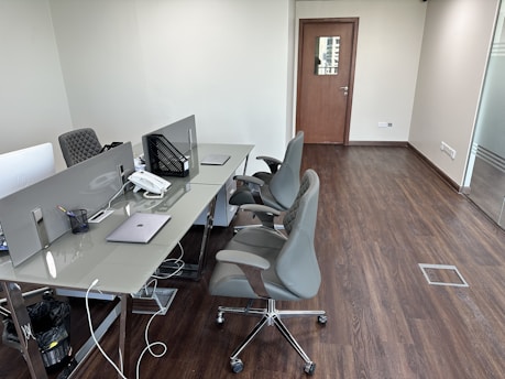 A modern office workspace with a row of desks equipped with dividers and ergonomic chairs. Items such as laptops, a phone, and stationery are neatly placed on the glass-topped desks. The office is minimally decorated, with wooden flooring and neutral-colored walls. A closed wooden door is focused in the background, and a floor-to-ceiling glass panel is seen on the right side.