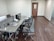 A modern office workspace with a row of desks equipped with dividers and ergonomic chairs. Items such as laptops, a phone, and stationery are neatly placed on the glass-topped desks. The office is minimally decorated, with wooden flooring and neutral-colored walls. A closed wooden door is focused in the background, and a floor-to-ceiling glass panel is seen on the right side.