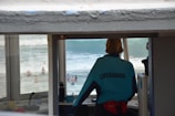 A lifeguard wearing a blue uniform stands inside a tower overlooking a beach area. The focus is on the lifeguard who is observing people swimming and playing in the ocean. The lifeguard’s jacket has the word 'LIFEGUARD' printed on the back, and the office is equipped with various items for monitoring the beach.