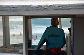 A lifeguard wearing a blue uniform stands inside a tower overlooking a beach area. The focus is on the lifeguard who is observing people swimming and playing in the ocean. The lifeguard’s jacket has the word 'LIFEGUARD' printed on the back, and the office is equipped with various items for monitoring the beach.
