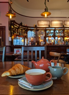 A cozy cafe setting featuring a wooden table with a croissant, a cup of tea on a saucer, a small milk jug, and a matching teapot. Elegant lighting fixtures hang from the ceiling, illuminating the warm and inviting atmosphere of the cafe, which has a well-stocked bar in the background.