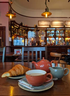 A cozy cafe setting featuring a wooden table with a croissant, a cup of tea on a saucer, a small milk jug, and a matching teapot. Elegant lighting fixtures hang from the ceiling, illuminating the warm and inviting atmosphere of the cafe, which has a well-stocked bar in the background.
