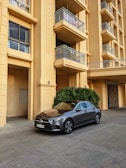 A modern, gray sedan is parked in front of a beige apartment building with multiple balconies. The building has plants at the entrance and features metal railings on the balconies. The car is positioned on a concrete pavement.