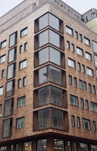A modern multi-story building with a distinctive facade made of wooden shingles. The architecture features large glass windows and balconies creating a geometric pattern. Various levels of the building are visible, showcasing a consistent design with repeated window arrangements.
